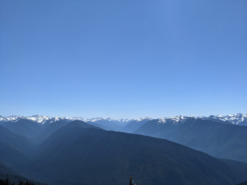 A view of several gracefully curved valleys receding into the distance, with snow-capped mountains towering above them in the far distance.